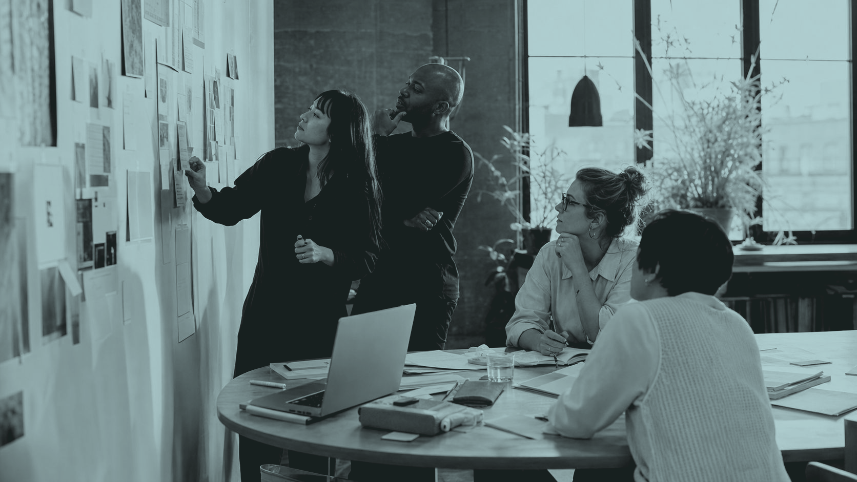 A team gathered around a table with laptops and printed briefs, discussing marketing work sketched across the wall behind them
